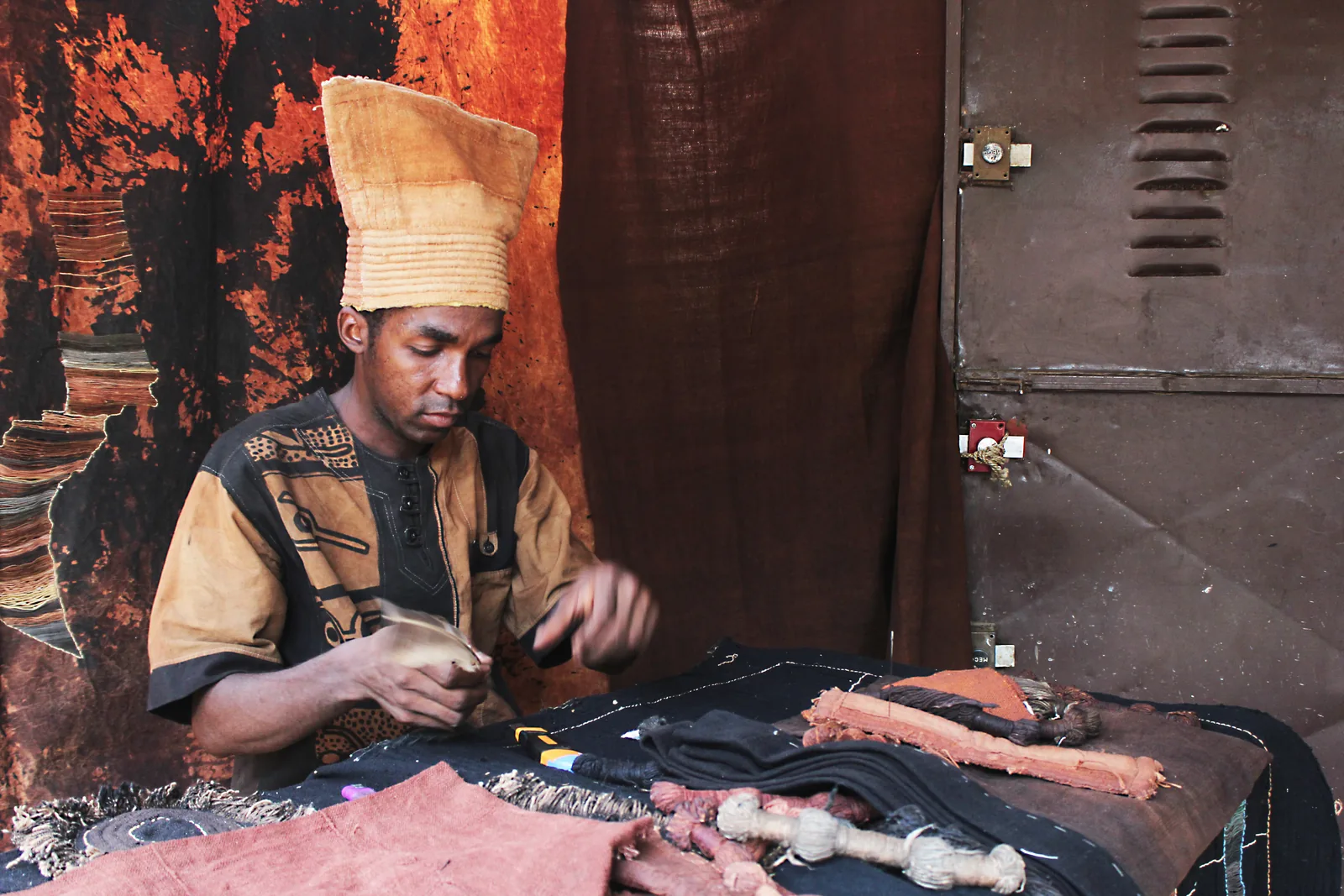 A man in traditional dress working on dark, patterned bogolan cloth at a table surrounded by finished textiles
