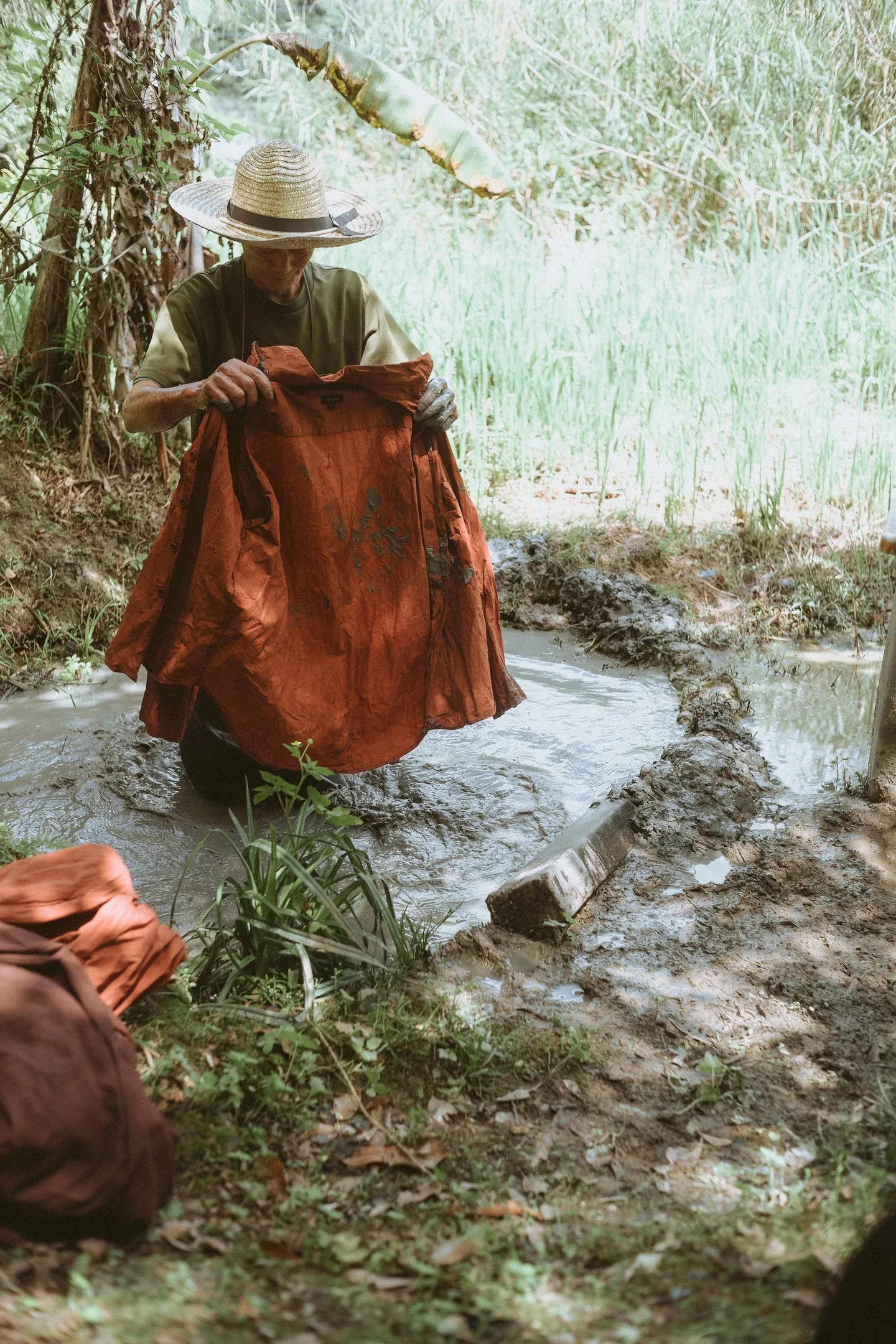 A worker in a straw hat kneeling in a mud paddy, holding up a rust-brown sharinbai-dyed shirt