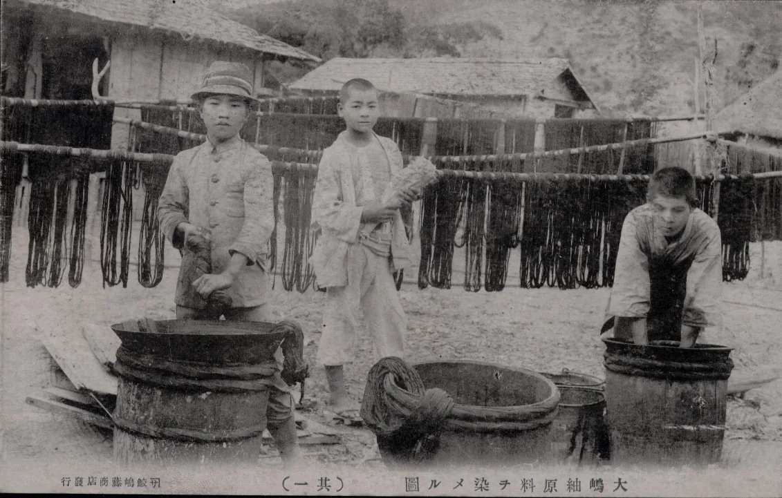 Black-and-white archival photograph of workers dyeing thread in barrels, with rows of dark dyed yarn hanks hanging to dry behind them