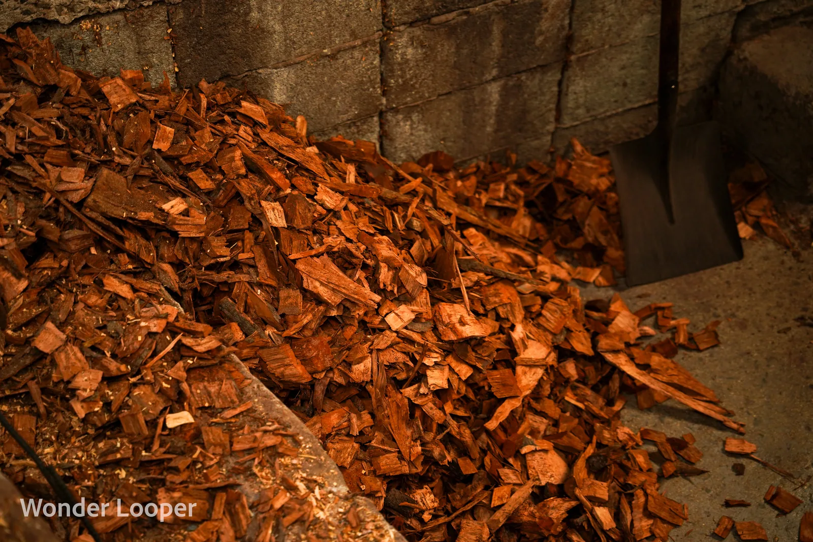 A large pile of reddish-brown wood chips on a concrete floor next to a stone block wall
