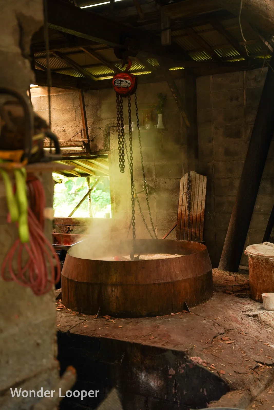 Steam rising from a large wooden dye kettle in a workshop, with an industrial chain hoist suspended above it