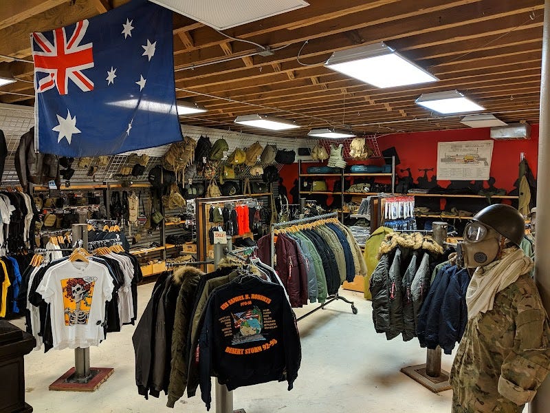 Basement level of Belmont Army Surplus in Chicago, with an Australian flag hanging overhead, dense racks of military jackets and graphic T-shirts, and a mannequin dressed in tactical gear and a gas mask.