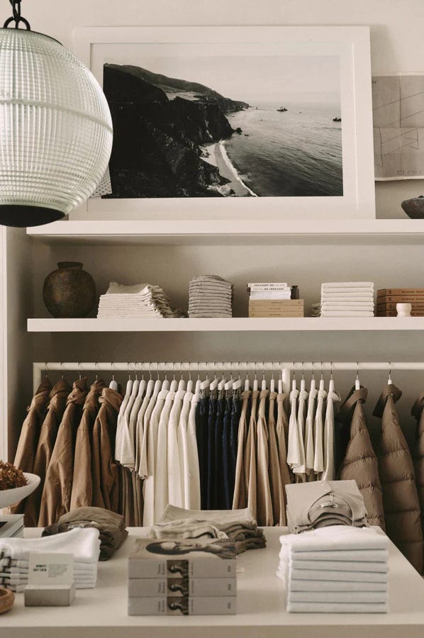 Minimalist white shelving display inside a Buck Mason store, with folded basics in neutral tones, stacked books, and a black-and-white coastal landscape photograph on the wall above.