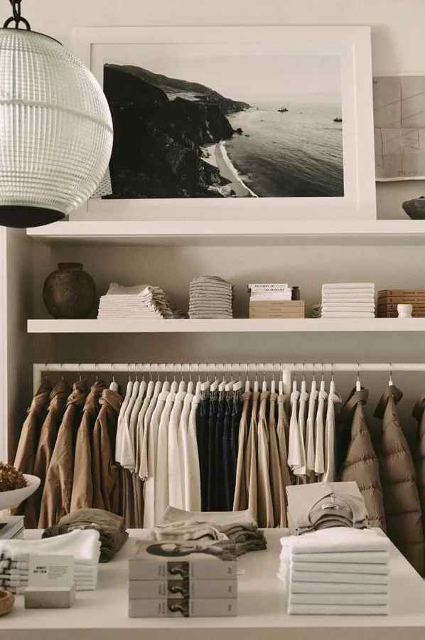 Minimalist white shelving display inside a Buck Mason store, with folded basics in neutral tones, stacked books, and a black-and-white coastal landscape photograph on the wall above.