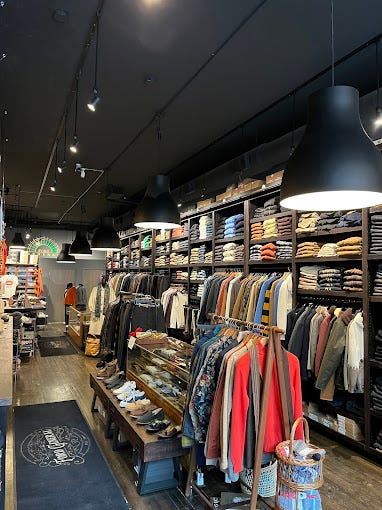 Interior of Ford General in Chicago, with dark walls and ceiling, densely packed wood shelving of folded shirts and jackets, and a patterned rug on the floor near the entrance.