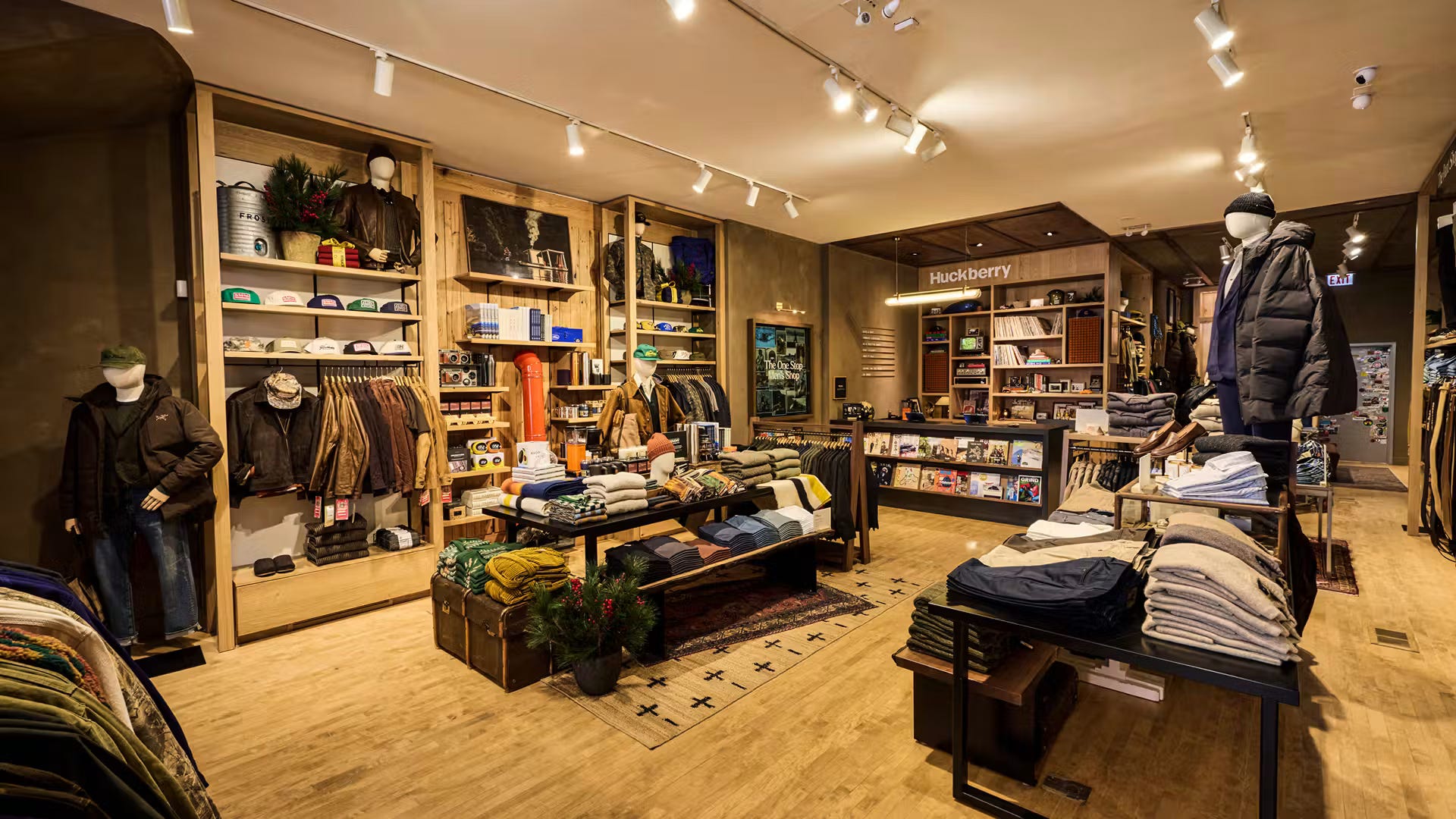 Wide view of the Huckberry Chicago store, with wood shelving holding caps, books, and folded garments, mannequins in outerwear, and a lit Huckberry sign at the back of the room.