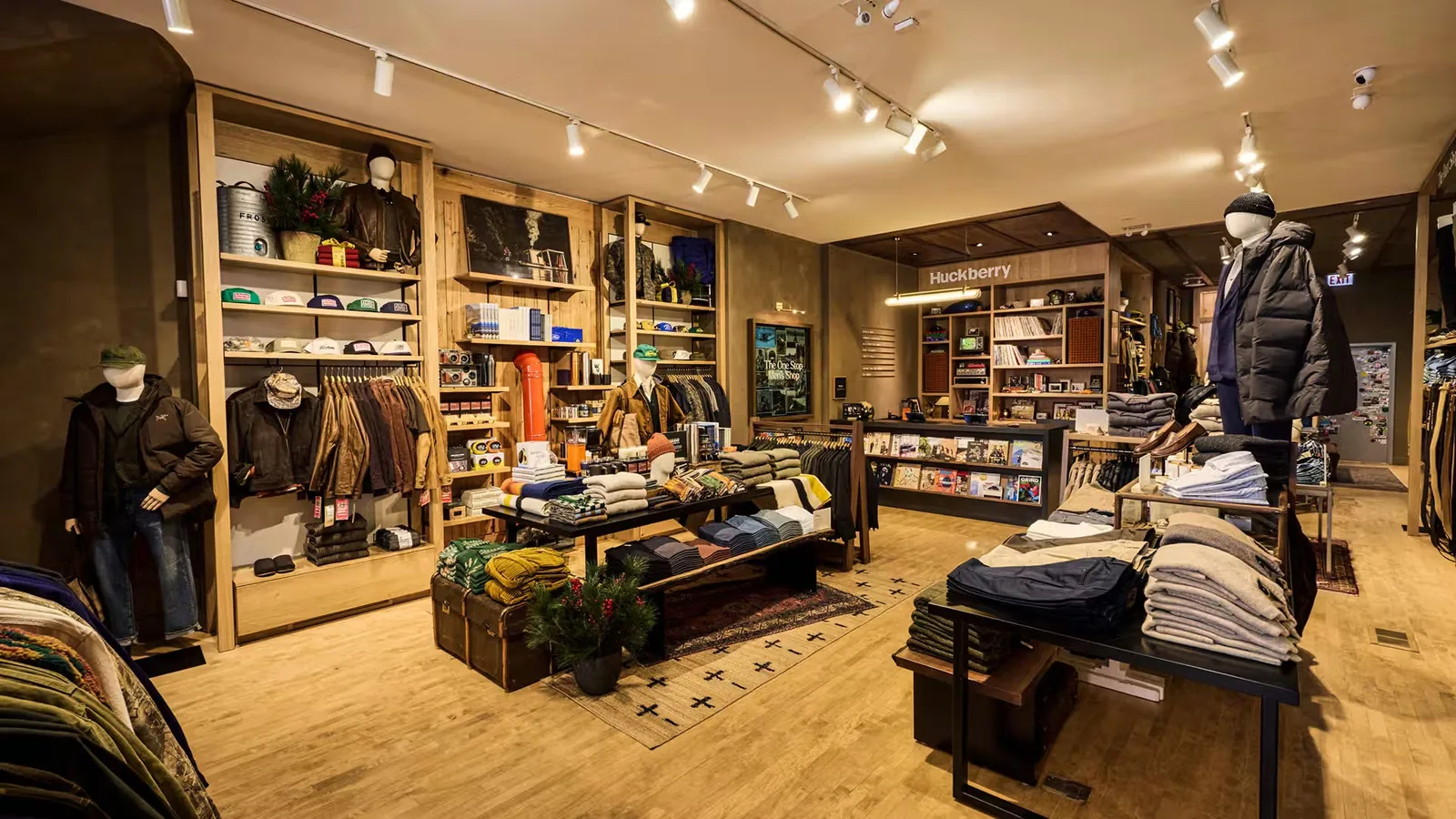Wide view of the Huckberry Chicago store, with wood shelving holding caps, books, and folded garments, mannequins in outerwear, and a lit Huckberry sign at the back of the room.