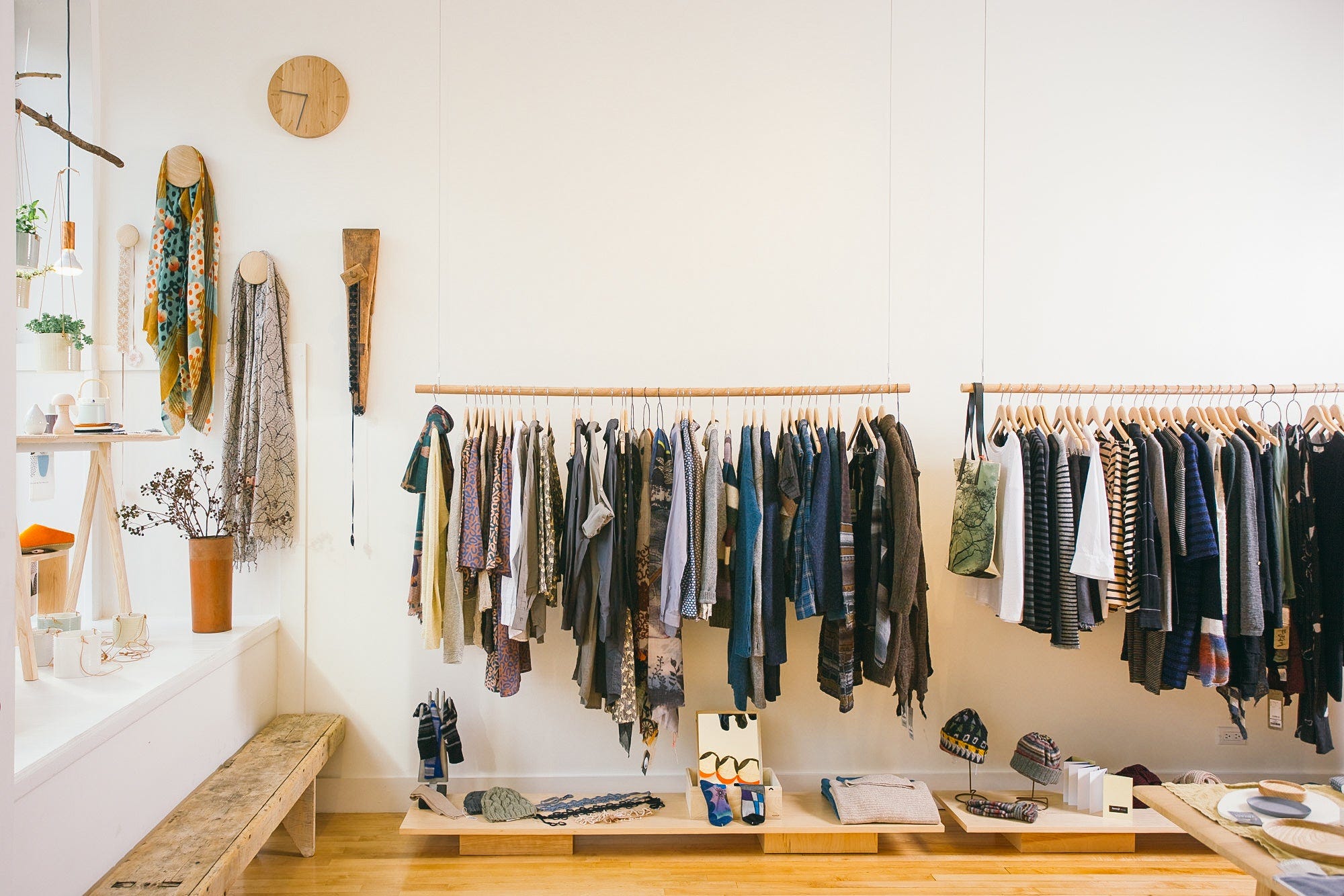 Bright, white-walled interior of Moth boutique in Chicago, with hanging scarves on the wall, a wooden clock, potted plants, and racks of sweaters and outerwear above tables of folded knitwear and hats.