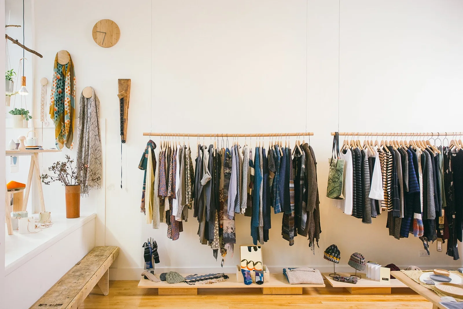 Bright, white-walled interior of Moth boutique in Chicago, with hanging scarves on the wall, a wooden clock, potted plants, and racks of sweaters and outerwear above tables of folded knitwear and hats.