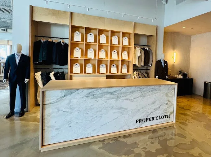Proper Cloth showroom in Chicago, with a marble-fronted reception desk, mannequins in tailored suits on either side, and shelves of folded dress shirts on the back wall.