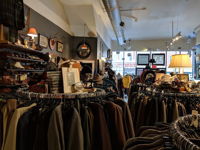 Densely packed interior of Richard's Fabulous Finds vintage shop, with racks of dark suits and sport coats, mounted taxidermy, framed portraits, and warm lamp lighting throughout.