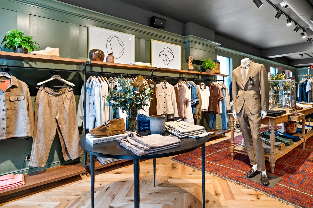 Todd Snyder Chicago retail floor with sage green walls, a tailored linen suit on a mannequin, racks of shirts and jackets, and a vintage rug on herringbone wood flooring.