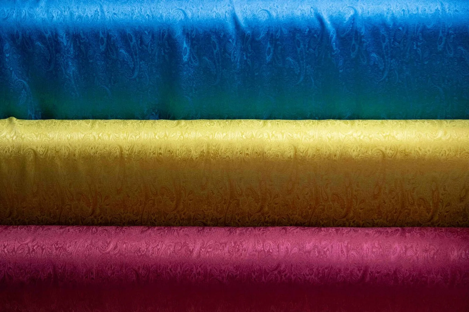 Three bolts of glossy, solid-colored fabric — blue, gold, and magenta — stacked in horizontal bands.