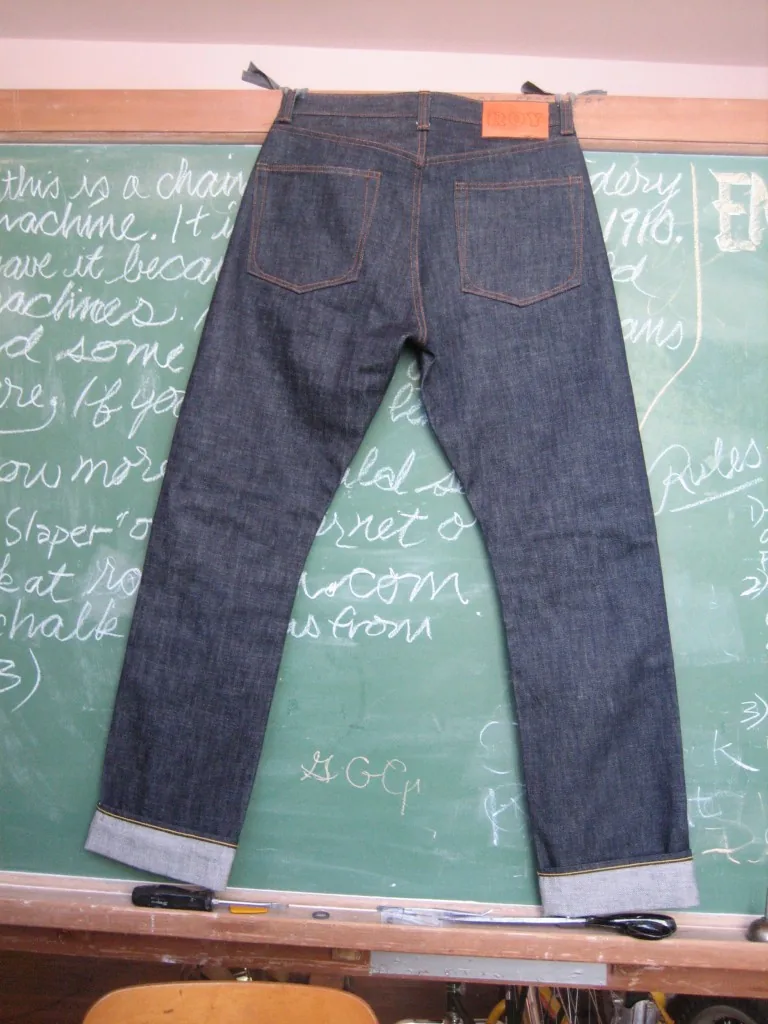 A pair of raw indigo selvedge jeans hung back-facing against a green chalkboard covered in handwritten notes about a 1910 chainstitch machine, cuffs turned up to show the selvedge ID.