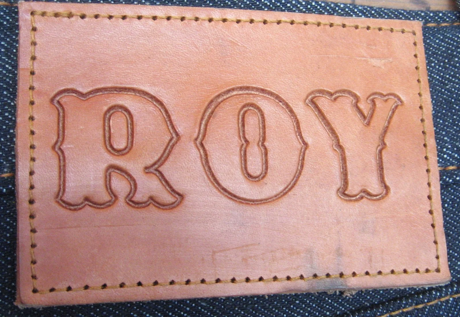 Close-up of a natural veg-tan leather patch embossed with the word ROY in outlined block letters, hand-stitched onto the waistband of raw indigo denim.