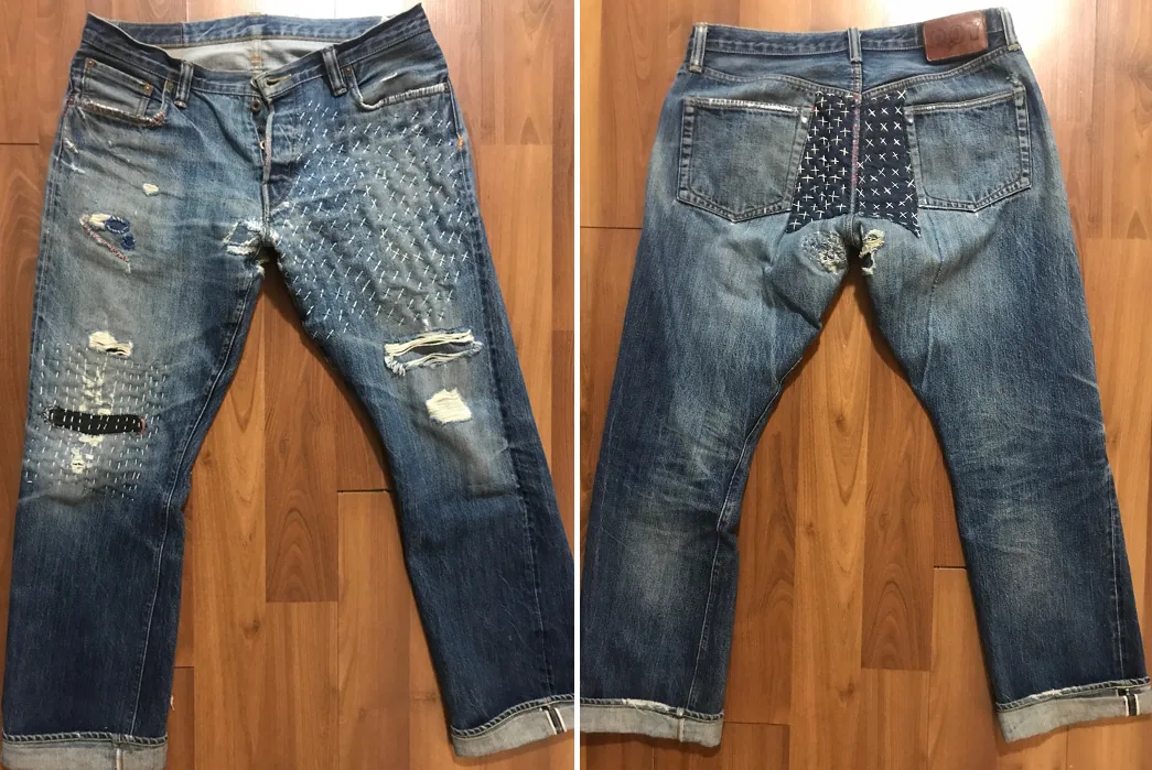 Front and back views of heavily faded selvedge jeans laid flat, with extensive white sashiko cross-stitch patching across one thigh and a dark indigo repair panel across the seat.