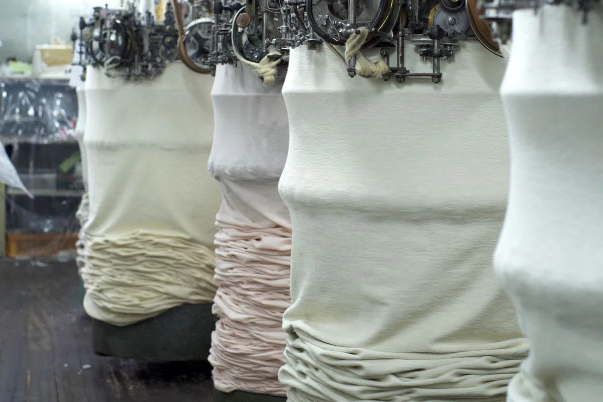 Vintage tsuriami loopwheel knitting machines with tubular knit fabric suspended beneath them.