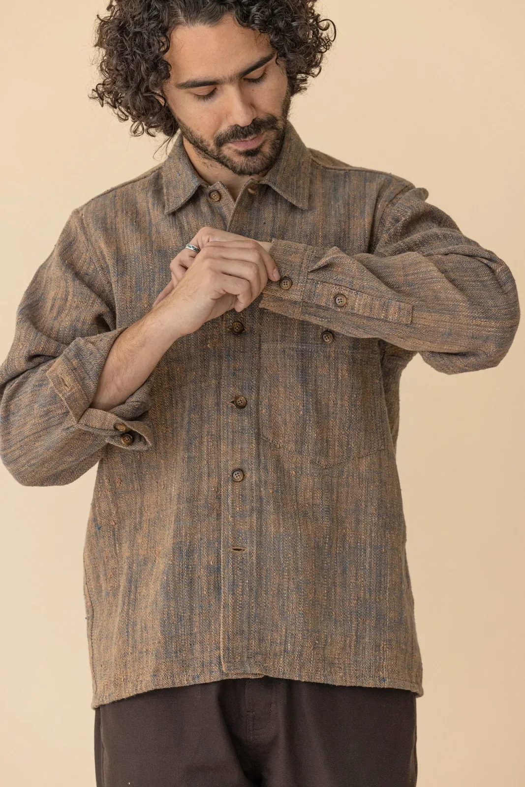 A studio product photo of a model buttoning the cuff of a hand-woven, earth-toned Graziano & Gutiérrez shirt over dark trousers.